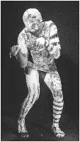 Description de l'image par IA : Homme en tenue de corps blanc et noir, bras croisés sur le torse, jambes légèrement écartées, sur fond sombre.
