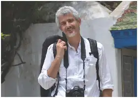 Description de l'image par IA : Homme avec appareil photo et sac à dos, souriant, devant un mur blanc et une maison bleue.