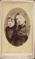 Description de l'image par IA : Deux jeunes filles posent ensemble, vêtues de robes ornées. L'une a les cheveux courts, l'autre longs.