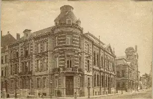 Description de l'image par IA : Grand bâtiment orné avec une tour ronde, fin XIXe siècle. Façade détaillée, fenêtres nombreuses, rue pavée devant.