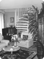 Description de l'image par IA : Salon avec canapé, table basse, fleurs, escalier, bibliothèque, plantes, et décoration.