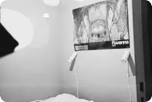 Description de l'image par IA : Image en noir et blanc d'une chambre avec un grand poster "Ravenna" sur le mur, deux lampes murales et un lit avec des draps blancs.
