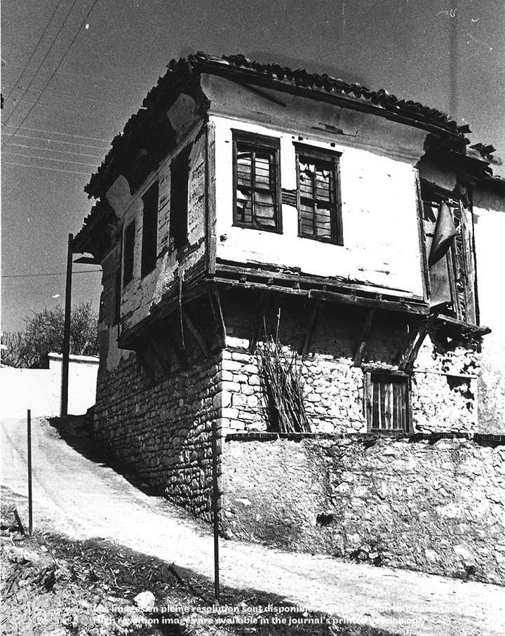 Image description generated by AI: Black and white photo of an old, two-story rural house with wooden shutters and a tiled roof, elevated on stone pillars.