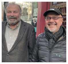 Description de l'image par IA : Deux hommes âgés, l'un avec une barbe et l'autre portant une veste noire, posent ensemble devant une boutique.