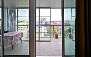 Description de l'image par IA : Fenêtre ouverte sur balcon avec table et plantes, vue sur immeubles et ville.