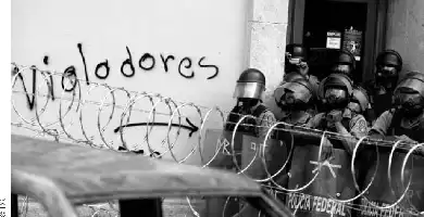 Description de l'image par IA : Des policiers en armure derrière une barrière de fil de fer barbelé devant un mur avec l'inscription "Vigiladores".