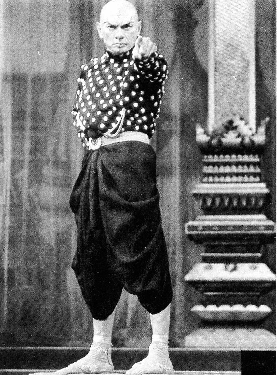 Description de l'image par IA : Homme chauve, vêtu d'un haut à pois et d'un pantalon large, debout, main sur le menton, devant un autel.