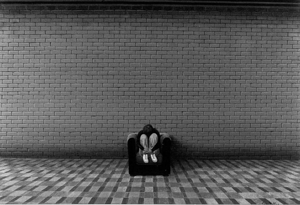 Description de l'image par IA : Un jeune homme assis dos à un mur de briques, les pieds sur un sol à damier.