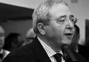 Description de l'image par IA : Homme âgé portant des lunettes, parlant dans un microphone.