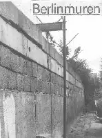 Description de l'image par IA : Mur de béton avec panneau "Berlinmuren" au-dessus, rue étroite à droite, arbres en arrière-plan.
