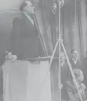 Description de l'image par IA : Homme en costume parle à un micro sur scène. À droite, un homme assis, probablement Léon Blum.