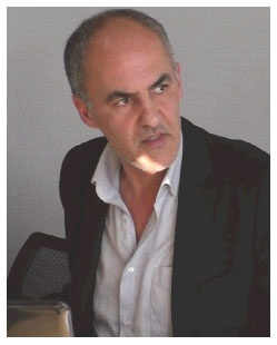 Description de l'image par IA : Homme âgé avec des cheveux gris, portant une veste noire et une chemise blanche, assis devant un micro.