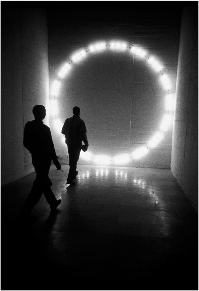 Description de l'image par IA : Deux silhouettes marchent dans une pièce faiblement éclairée avec un grand cercle lumineux au fond.
