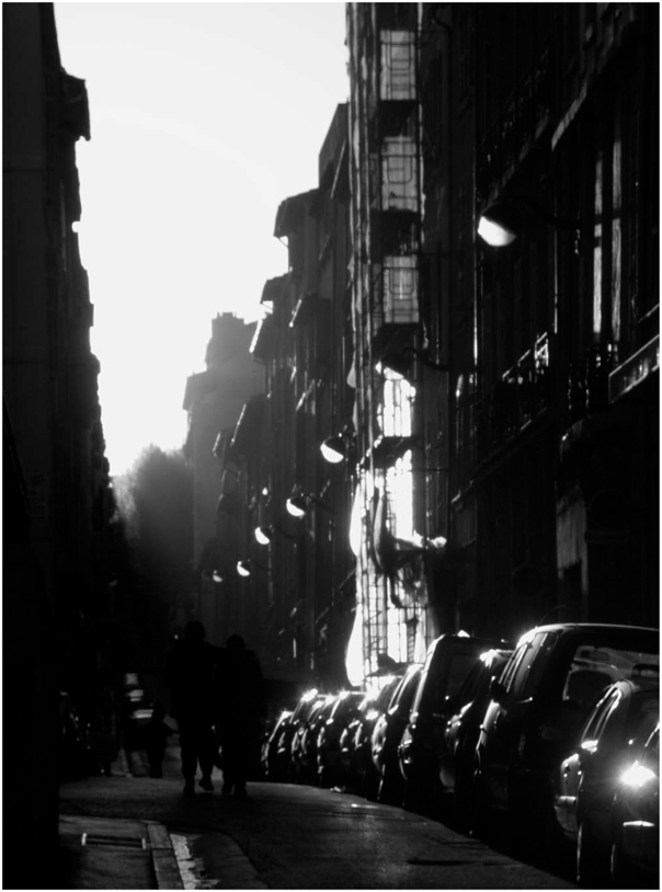 Description de l'image par IA : Rue étroite avec voitures garées, bâtiments hauts, quelques piétons.