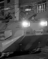 Description de l'image par IA : Voiture blindée avec deux phares puissants, stationnée dans une rue étroite avec des bâtiments et des fils électriques en arrière-plan.