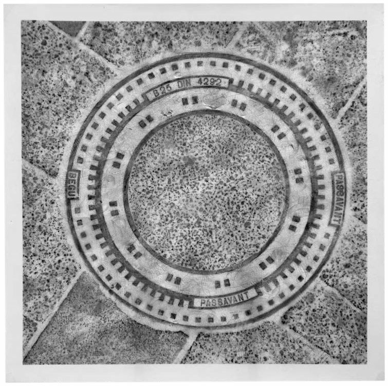 Description de l'image par IA : Plaque circulaire avec inscriptions et motifs gravés sur sol en dalles.