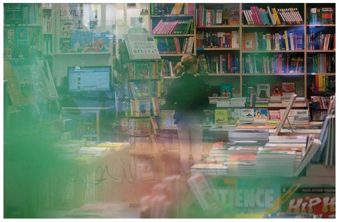 Description de l'image par IA : Bibliothèque avec des étagères remplies de livres, une personne travaillant derrière un comptoir, ordinateur et livres exposés.