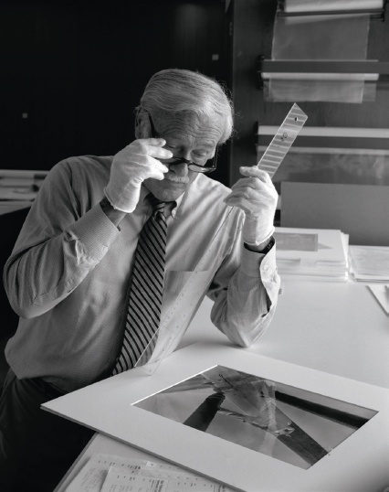 Description de l'image par IA : Homme âgé avec lunettes, tenant une règle et regardant une photo en noir et blanc sur une table.