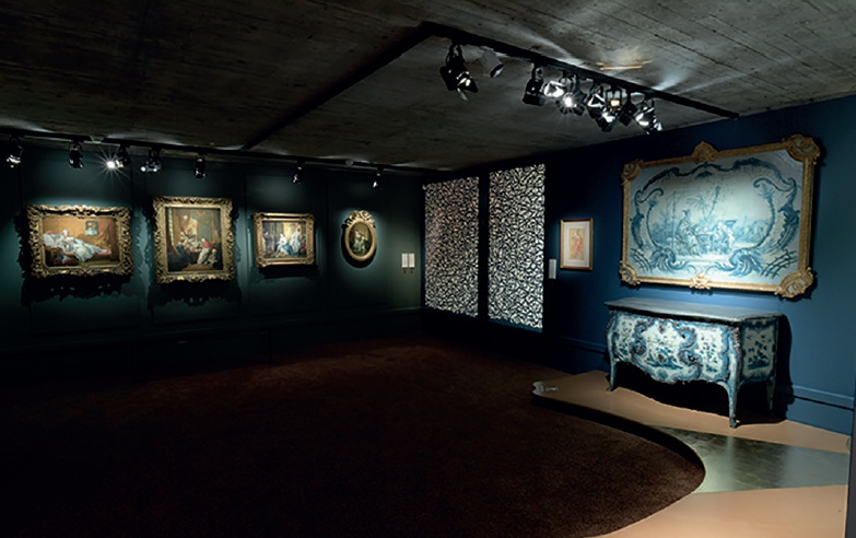 Description de l'image par IA : Salle d'exposition avec des tableaux et meubles, murs bleus, éclairage ciblé.