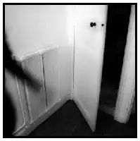 Description de l'image par IA : Une porte blanche ouverte révélant une pièce sombre avec un radiateur et un rideau blanc.