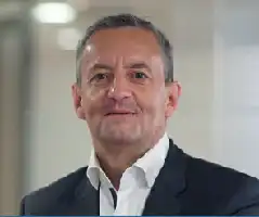 Description de l'image par IA : Homme d'âge moyen avec des cheveux gris, portant une veste et une chemise, souriant dans un cadre intérieur.