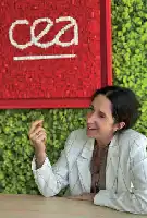 Description de l'image par IA : Femme en veste blanche devant un mur rouge avec le logo CEA et une verdure, elle sourit et pointe du doigt.
