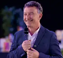 Description de l'image par IA : Homme souriant tenant un microphone, portant un costume bleu, en extérieur.