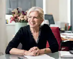 Description de l'image par IA : Femme âgée souriante, assise à un bureau avec des papiers, des fleurs et un ordinateur portable.