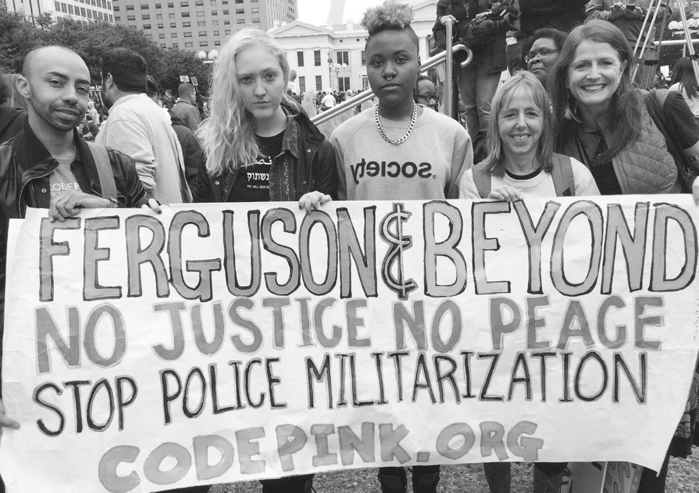 Description de l'image par IA : Manifestation avec banderole "Ferguson & Beyond, Justice, Pas de Paix, Arrêtez la Militarisation Policière".