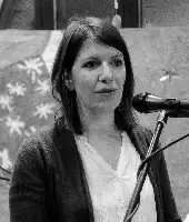 Description de l'image par IA : Femme parlant au microphone, cheveux mi-longs, veste et chemise blanche, fond avec motifs floraux.
