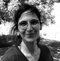 Description de l'image par IA : Femme aux cheveux longs et lunettes rondes, souriante, en noir et blanc, dehors avec des arbres en arrière-plan.