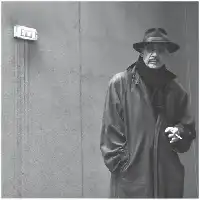 Description de l'image par IA : Homme en manteau et chapeau, cigarette à la main, debout contre un mur avec une plaque.