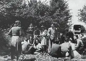 Description de l'image par IA : Groupe d'étudiants en plein air, certains assis, d'autres debout, entourés d'arbres et de livres.